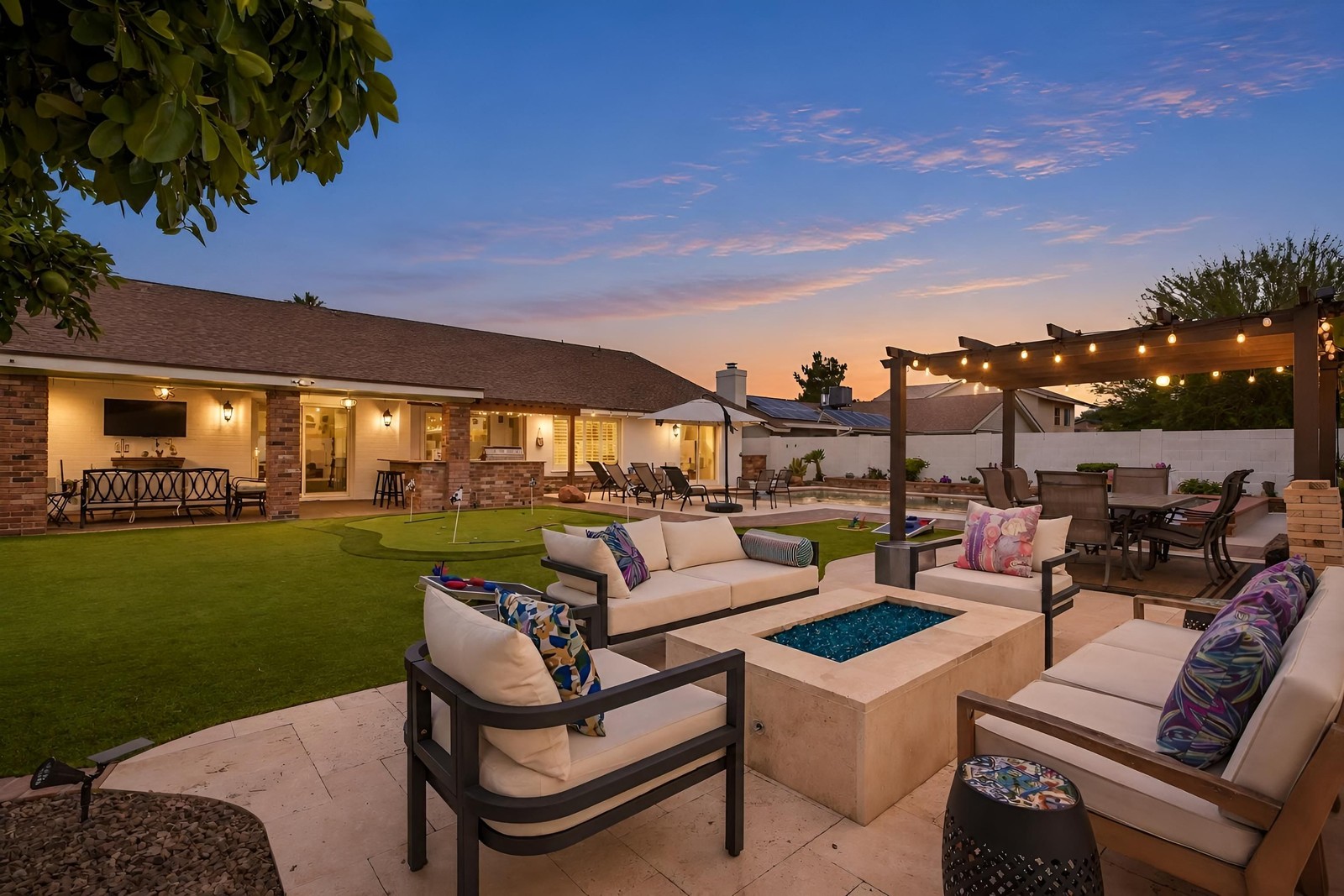 The same backyard as a virtual twilight: fire pit lit, pergola string lights on, sunset sky